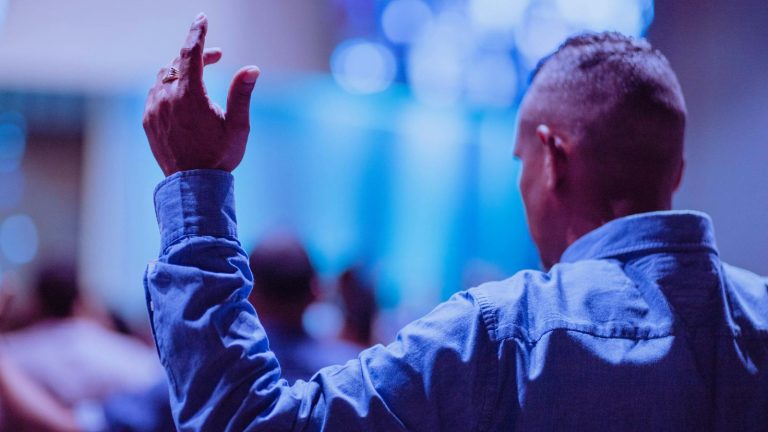 Back view of a man raising hand in prayer at a church service indoors.