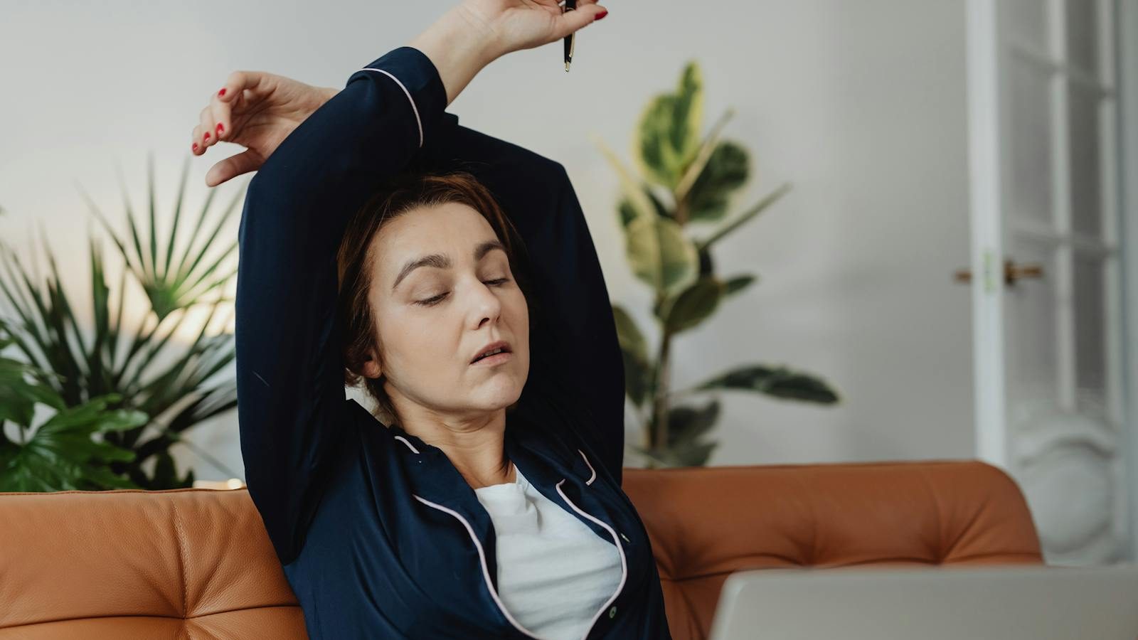 A tired woman in pajamas stretches while working remotely from her living room couch.