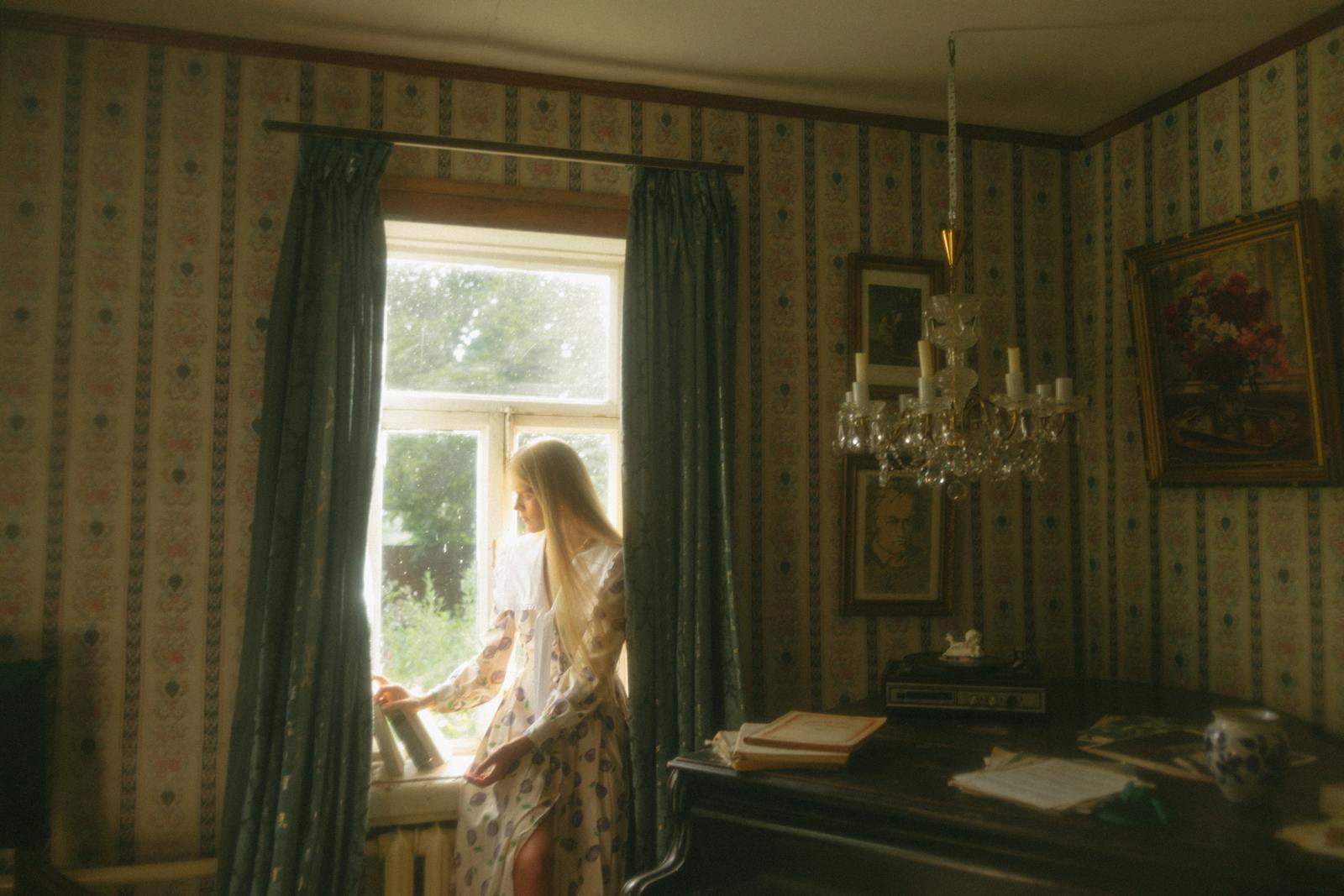 A young woman in a vintage room looking out a window, styled elegantly.