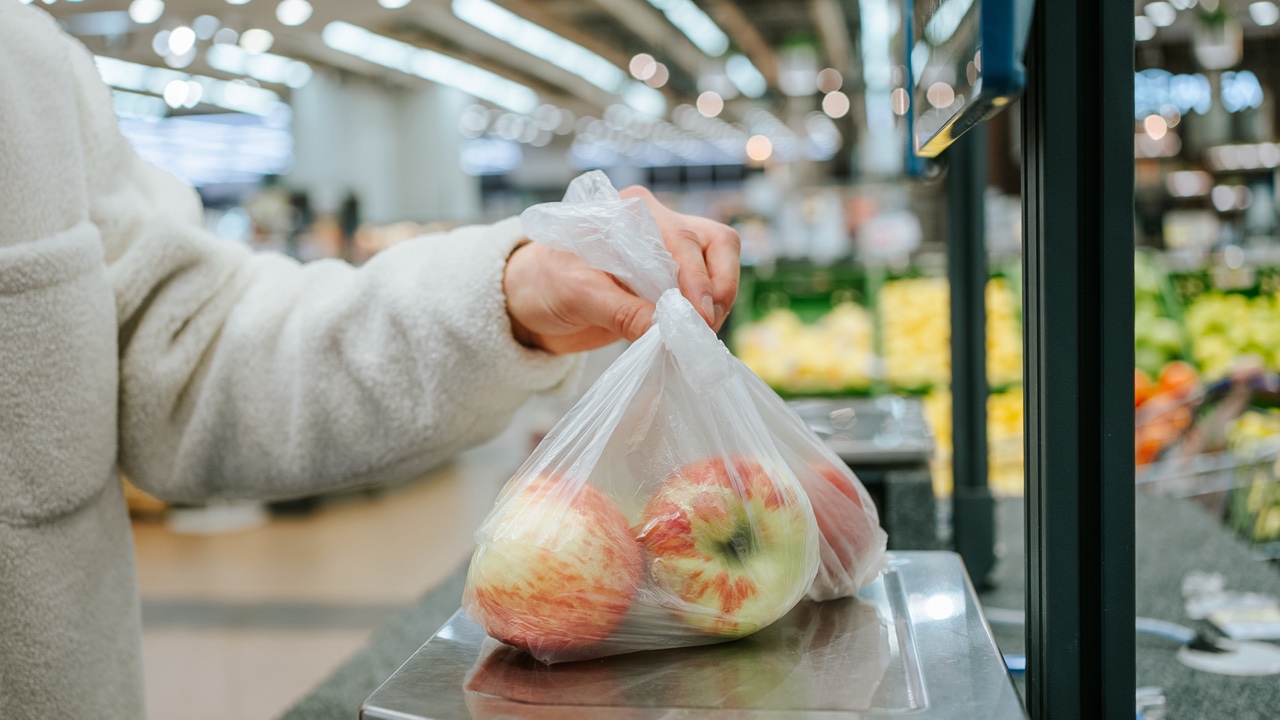A person's hand is shown placing a plastic bag of fresh apples on a digital scale in the produce section of a grocery store, ensuring the weight is correct for checkout