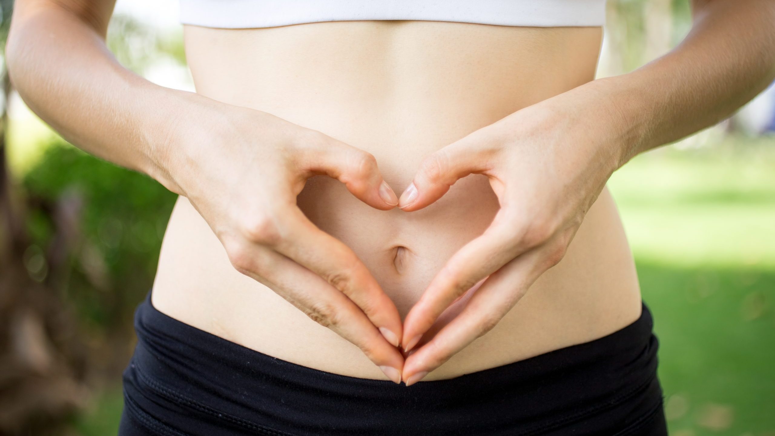 Close-up of female hands shaping heart on belly outdoors. Fitness and dieting concept