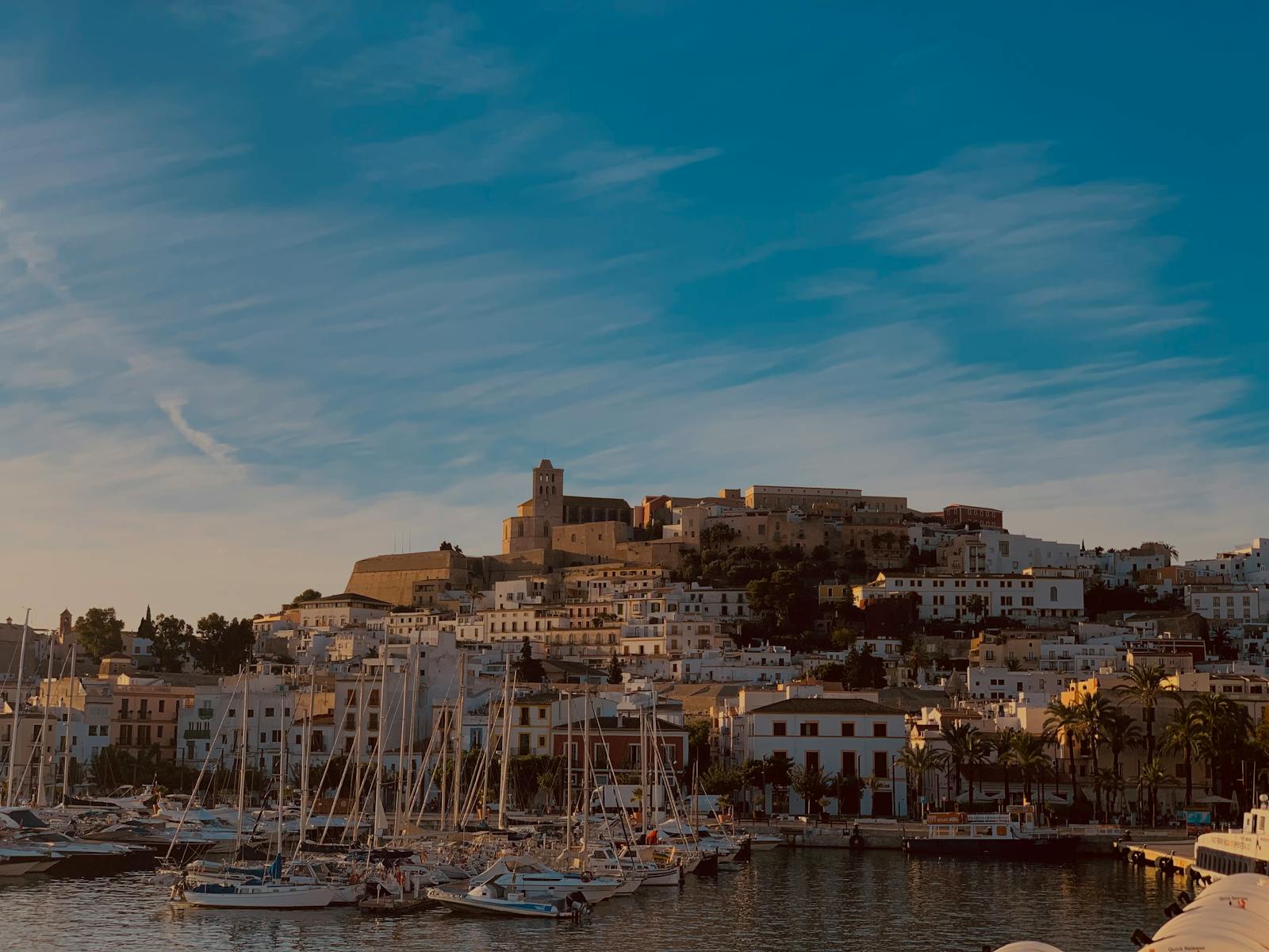 A tranquil view of Ibiza harbor with sailboats and historic buildings at dusk.