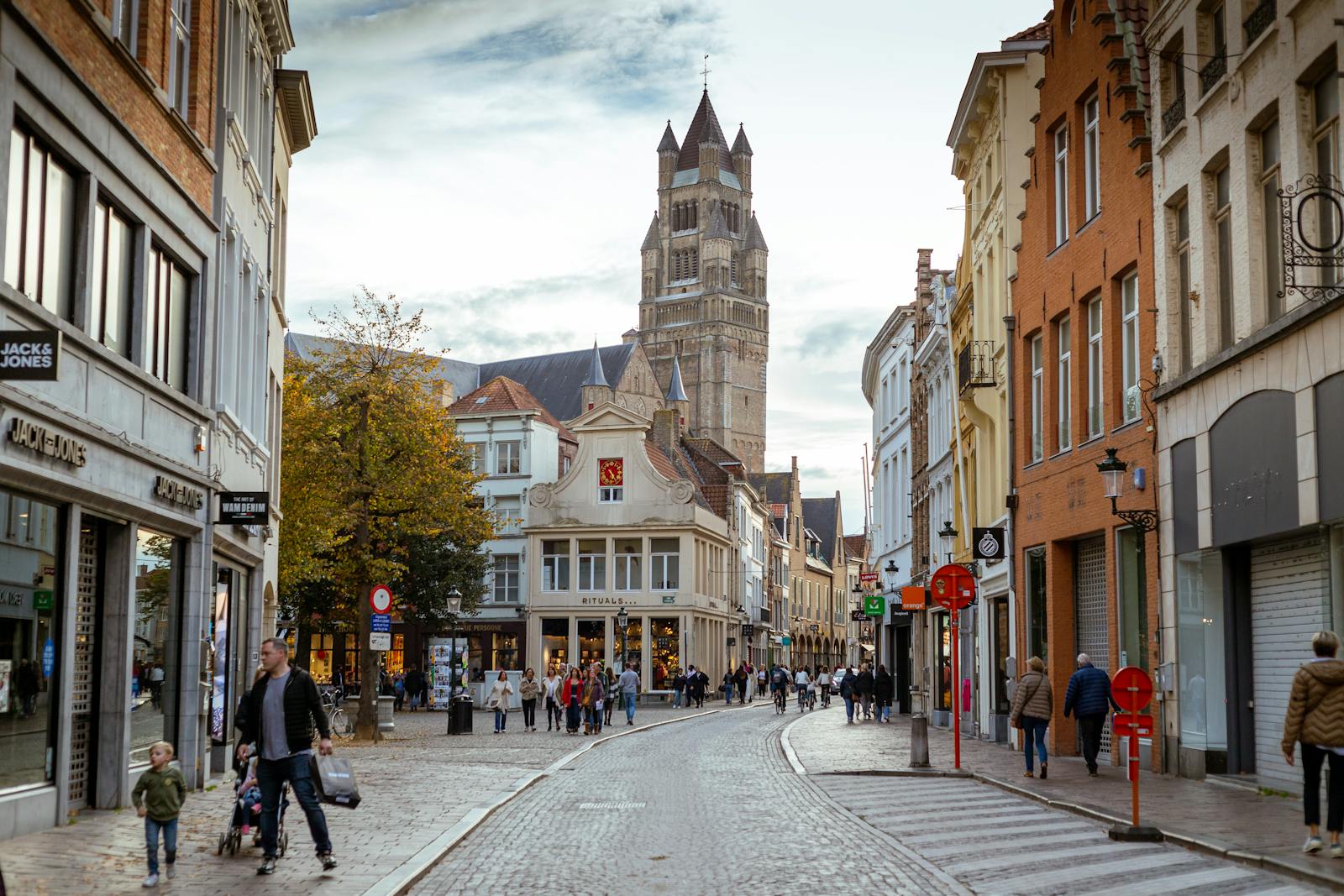 Picturesque street in Bruges with Belfry tower, cobblestones, and people strolling in autumn.