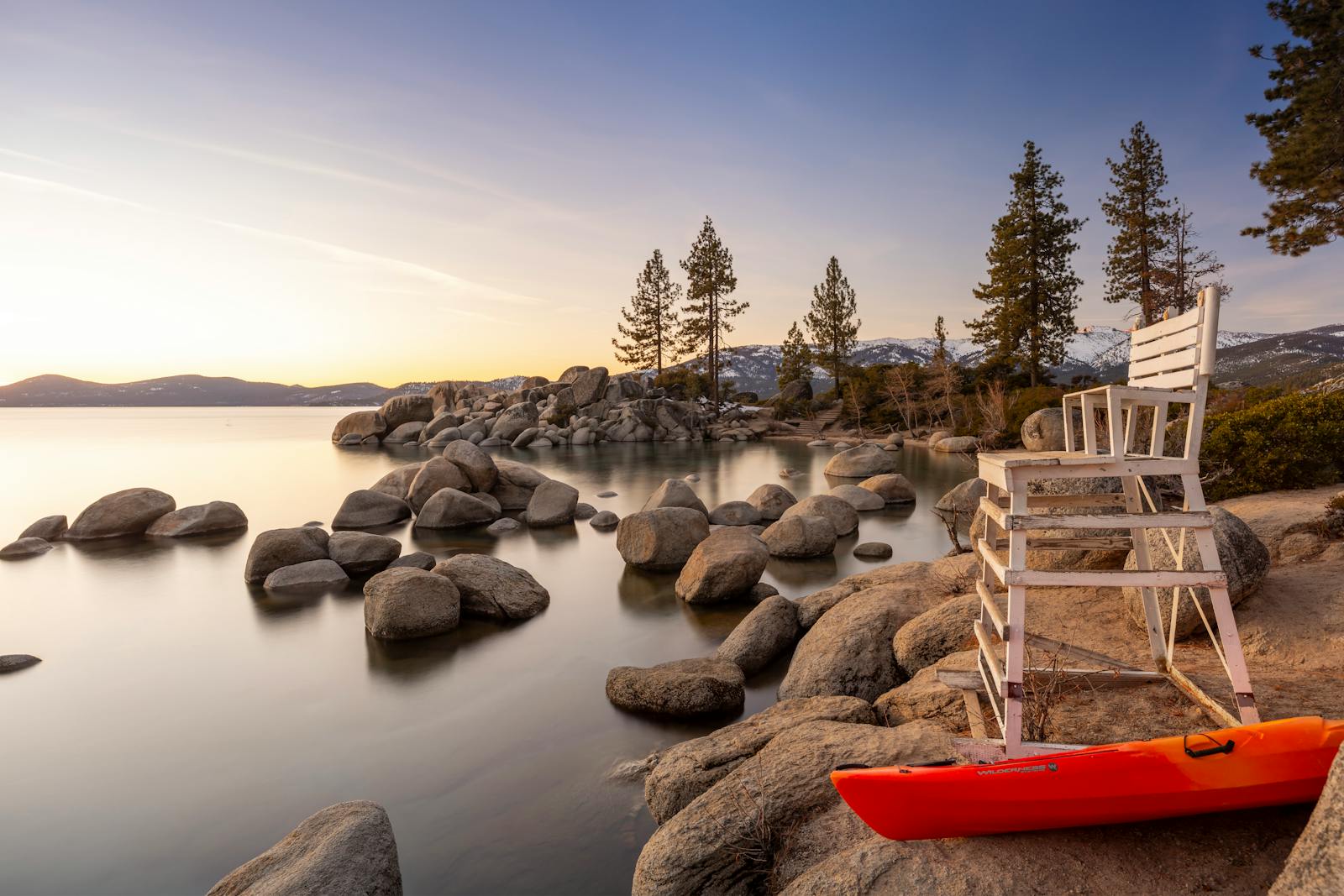 Golden sunset illuminating Lake Tahoe's serene waters, framed by rocks, trees, and a lifeguard chair.