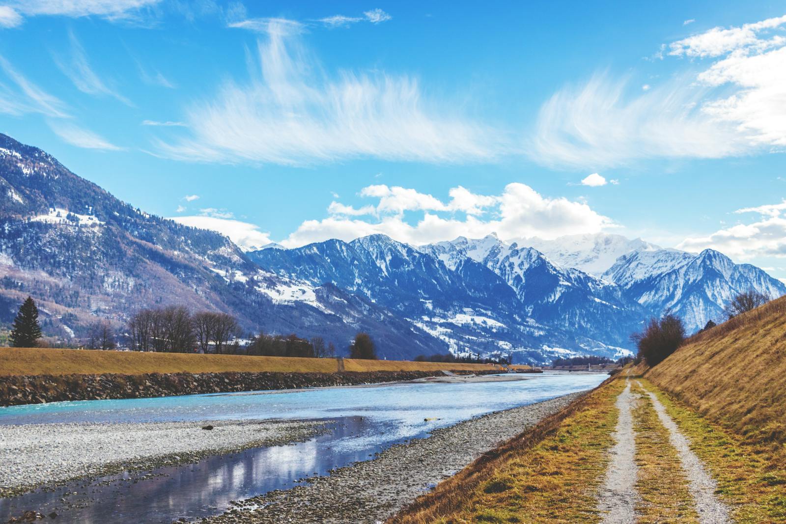 Explore the stunning Swiss Alps with a serene pathway alongside a flowing river under clear blue skies.