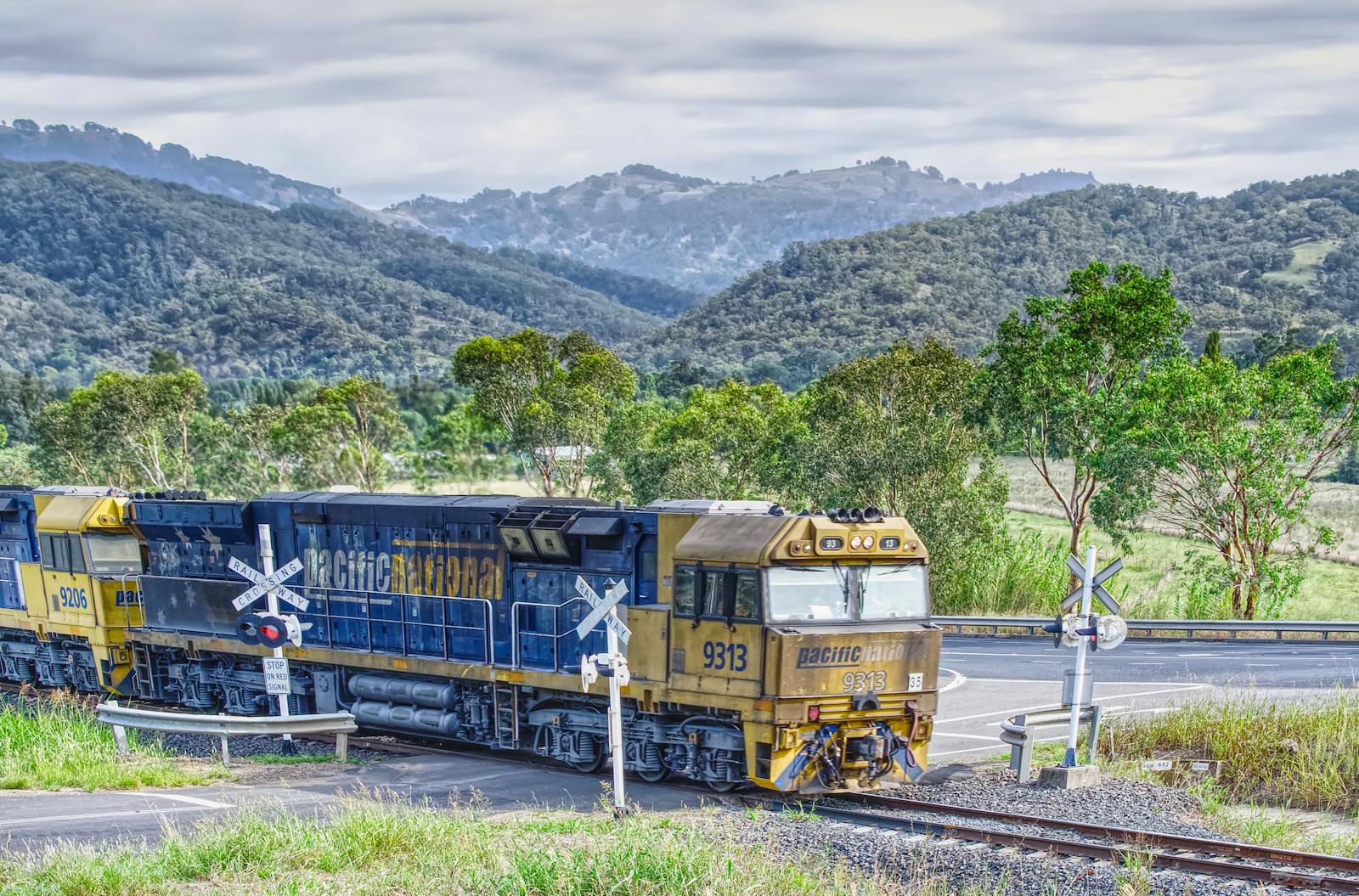 A freight train travels through a picturesque mountain landscape, crossing tracks amidst lush greenery.