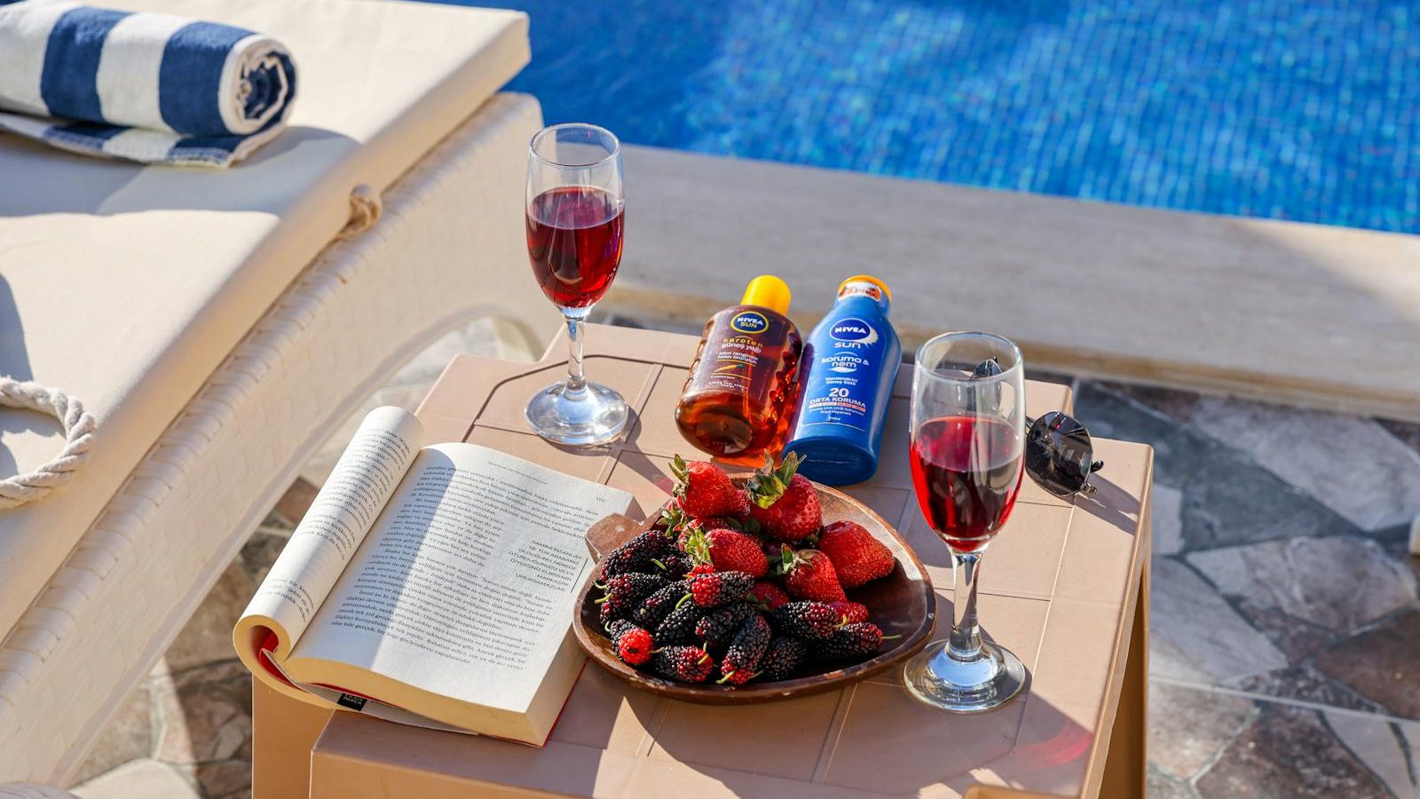 A beautiful poolside setup with red wine, fresh berries, and a book, perfect for relaxation.