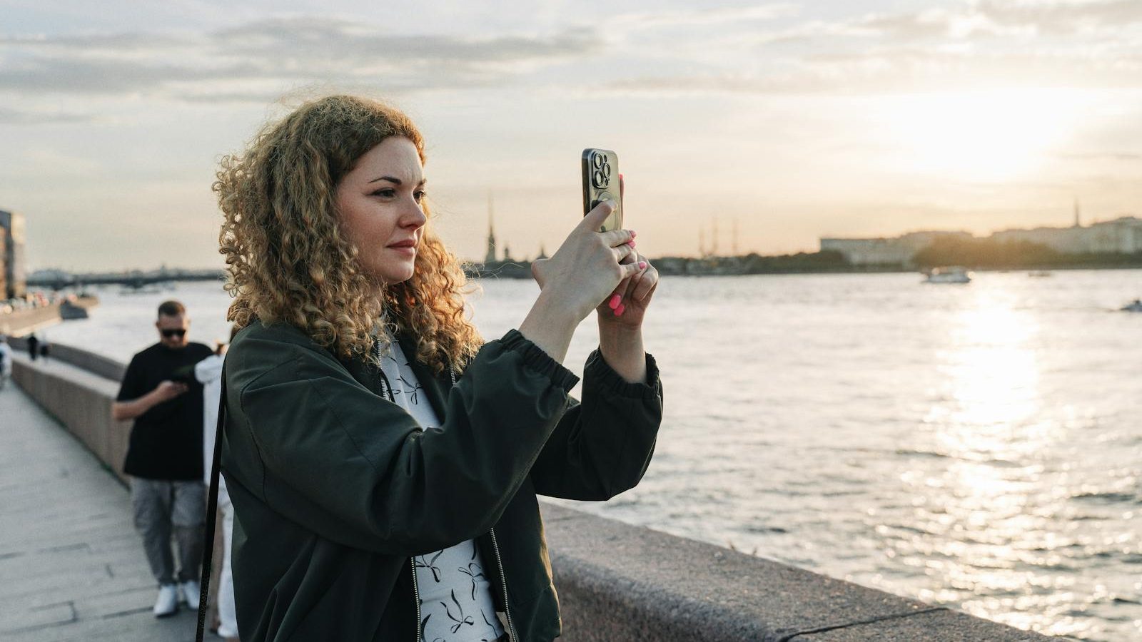 A woman captures sunset on her smartphone by the waterfront with scenic views.