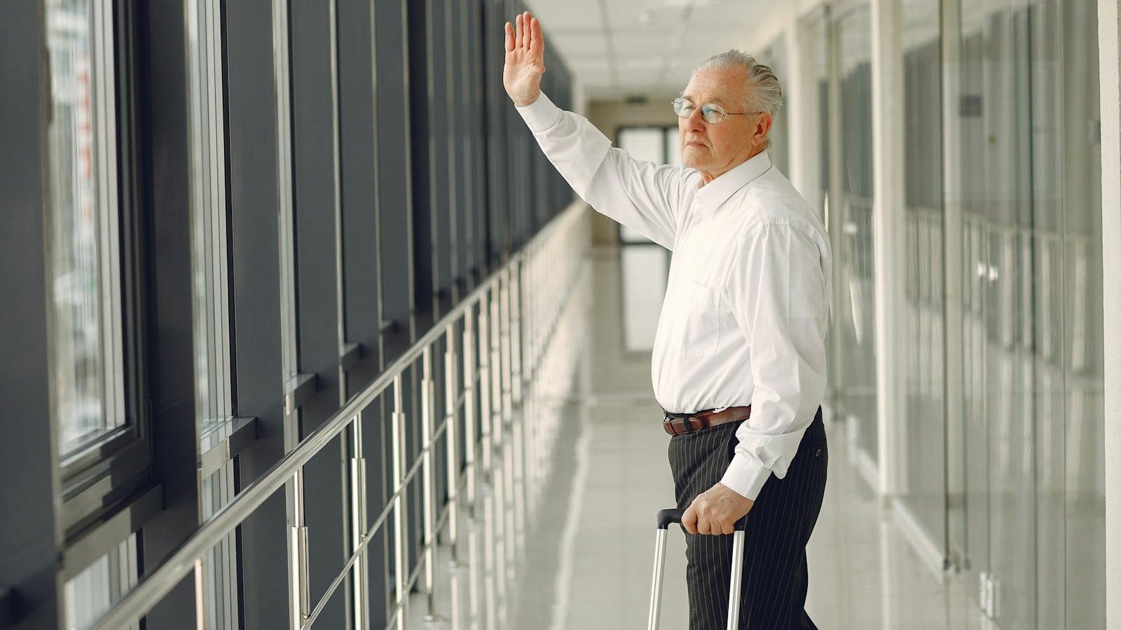 Side view of aged calm male in formal clothes with suitcase walking along modern airport hallway and waving goodbye while looking out glass wall