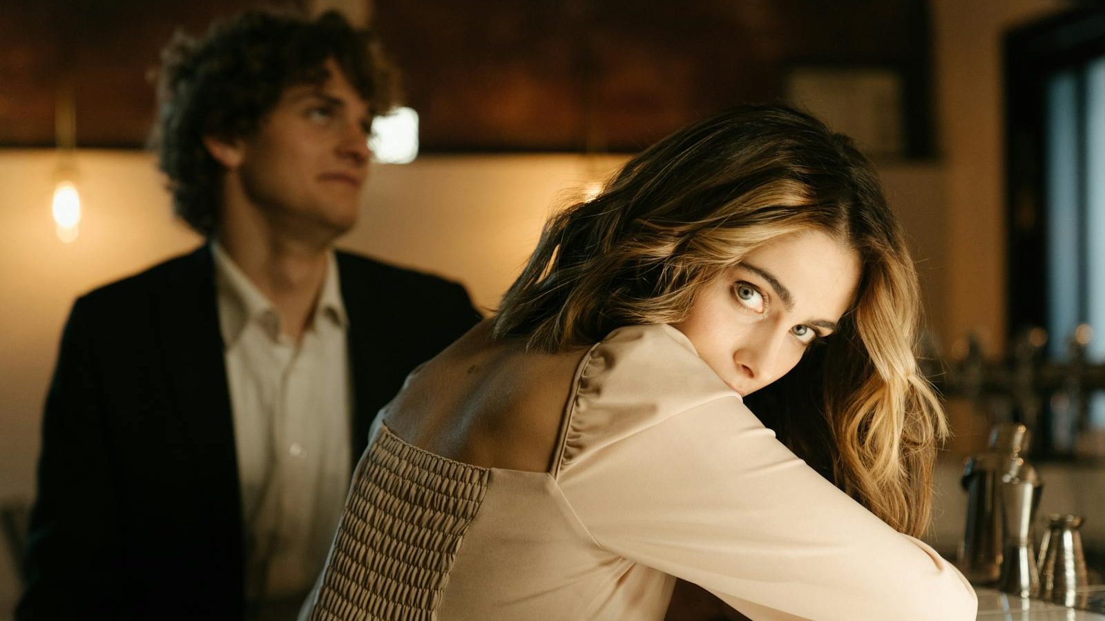 A stylish man and woman share a moment at a bar, with warm ambient lighting.