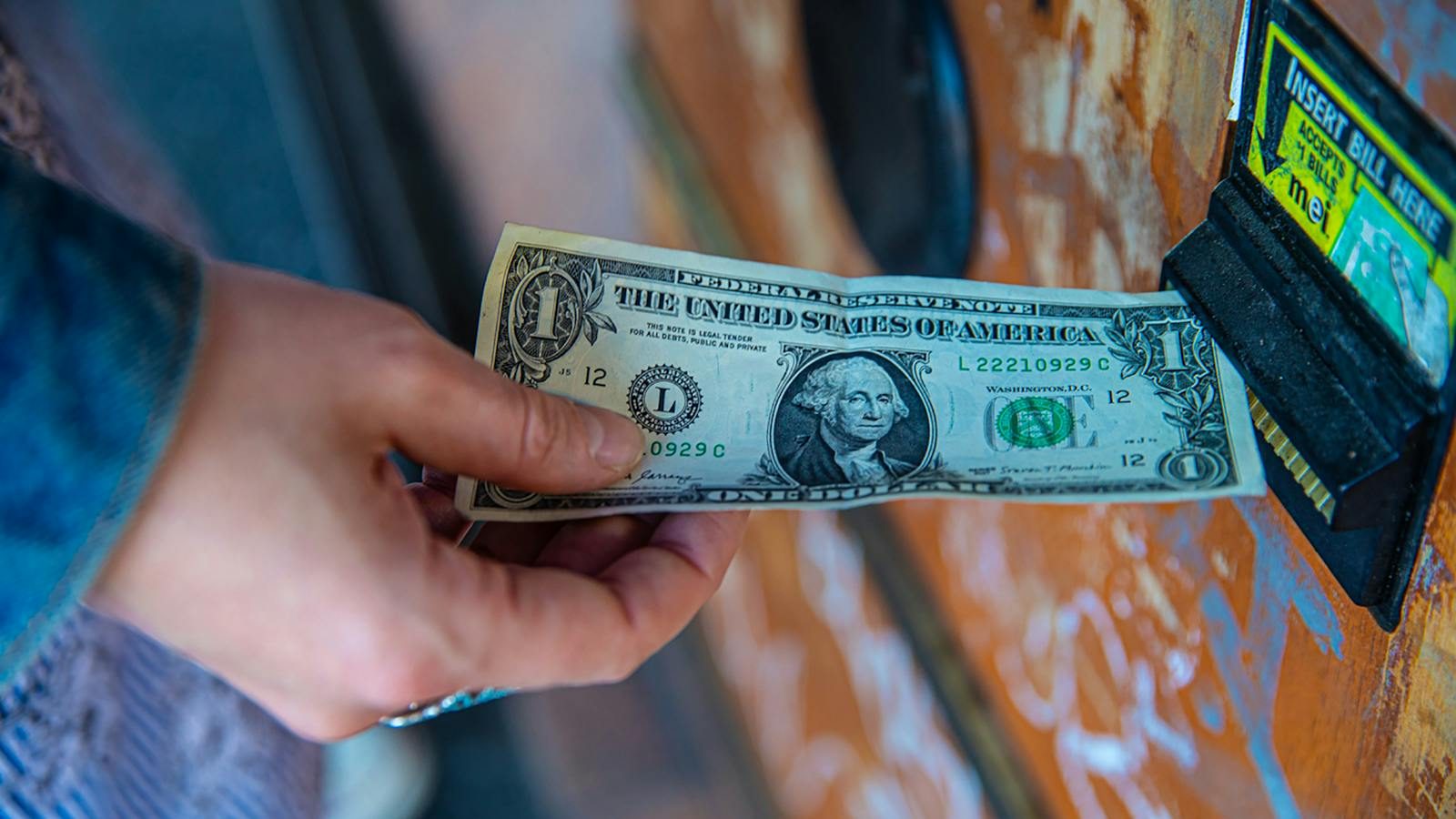 A person inserting a US dollar bill into a vending machine slot, capturing a financial transaction moment.
