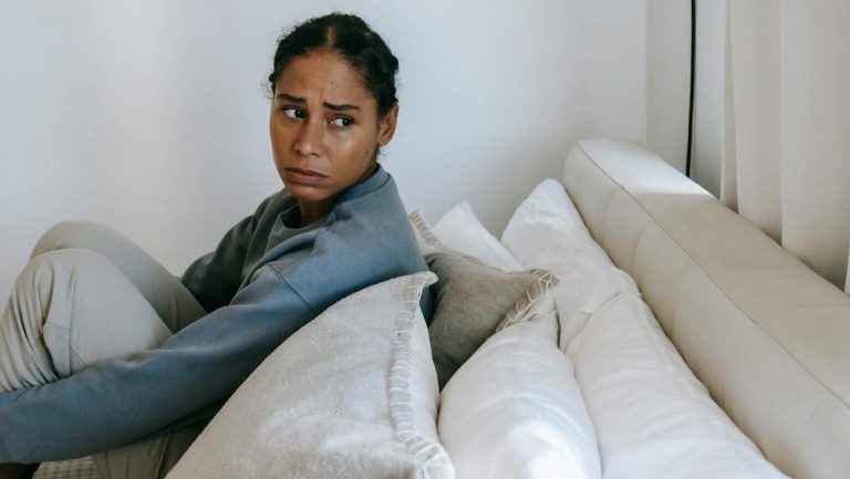 Sad young African American woman spending time on white bed and thinking in casual outfit while looking away at home