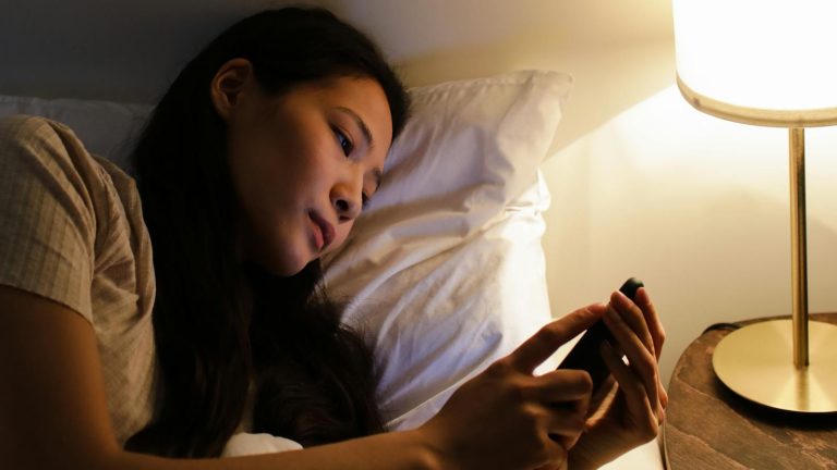 A woman lying in bed at night using a smartphone, illuminated by a bedside lamp.