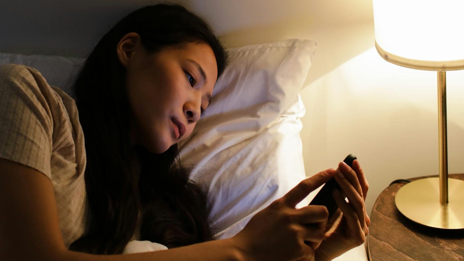 A woman lying in bed at night using a smartphone, illuminated by a bedside lamp.