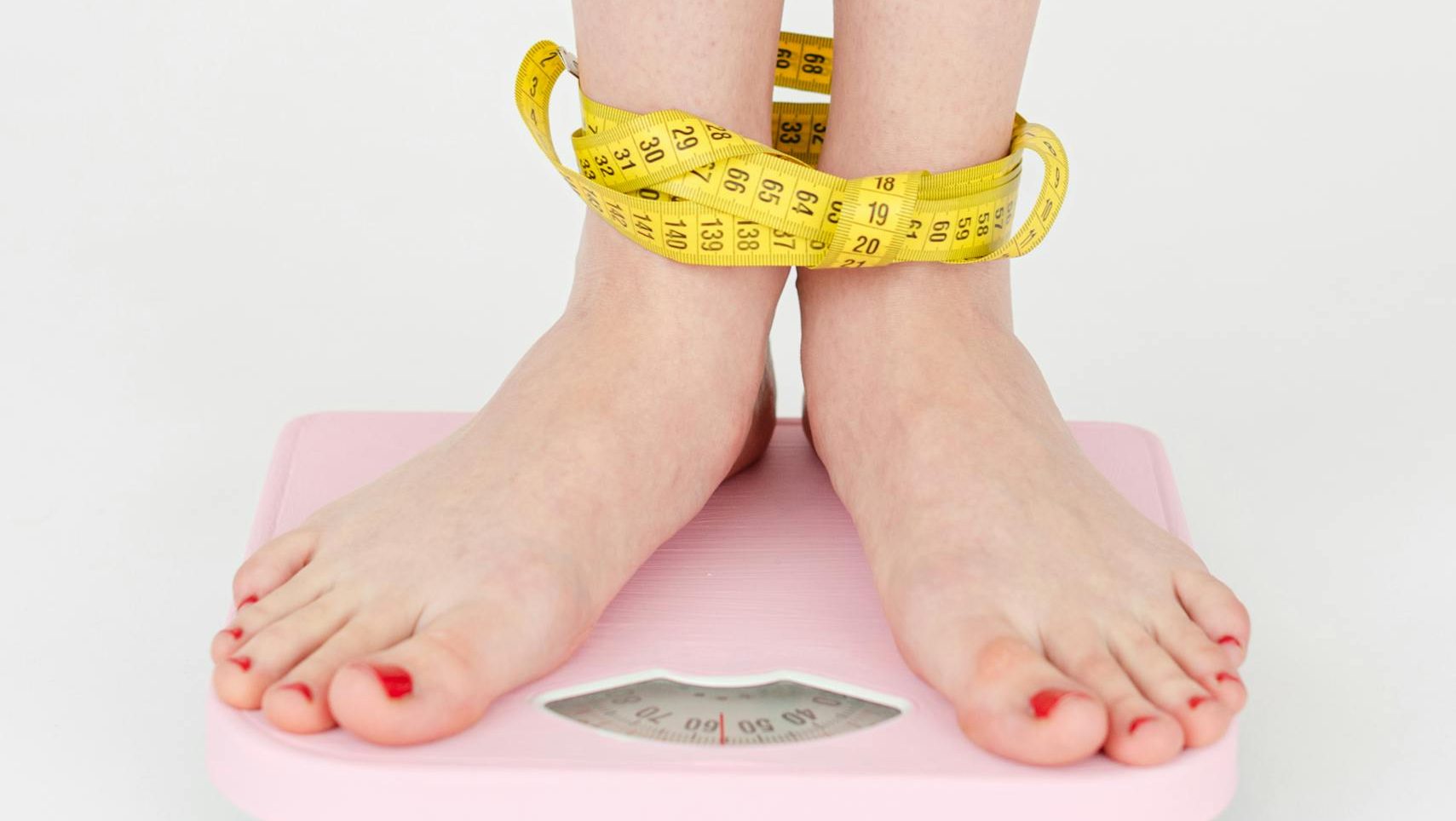 High angle of crop anonymous female with centimeter on legs checking weight on scales on white background