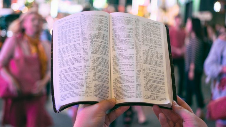 person holding bible on road with people walking on sidewalk beside buildings during nighttime