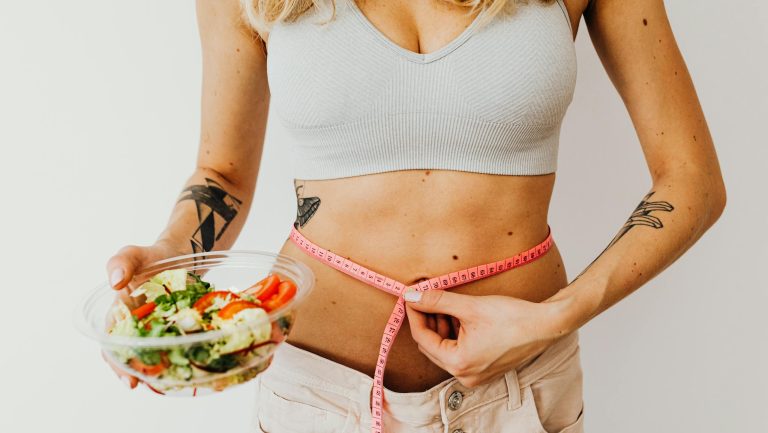 Woman measuring waistline while holding a fresh vegetable salad, emphasizing healthy living.