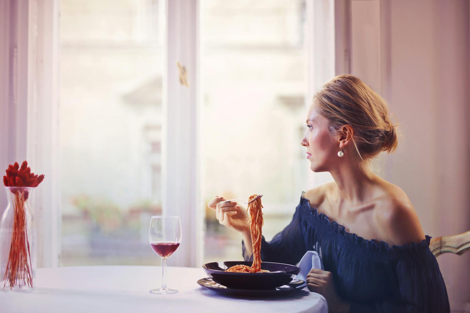 A woman enjoying a meal of spaghetti and wine at a restaurant table, elegantly dressed.