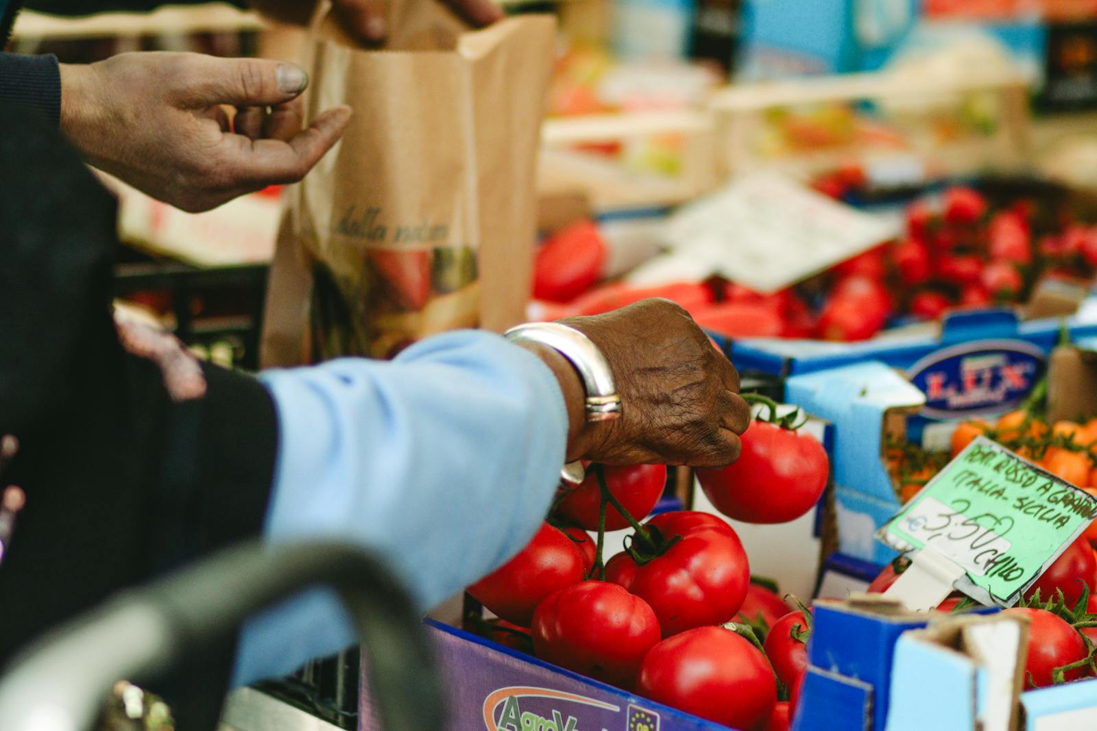 Close-up of elderly hands selecting ripe tomatoes at an outdoor vegetable market.