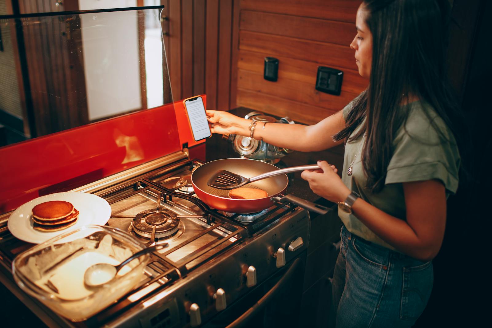 A woman uses a phone recipe to cook pancakes on a stove in a modern kitchen.