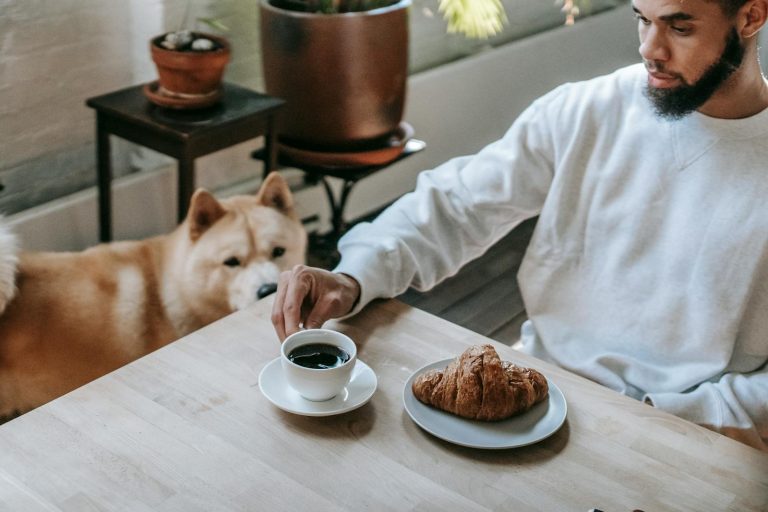 A man enjoying breakfast with coffee and croissant, accompanied by his Akita Inu dog at home.