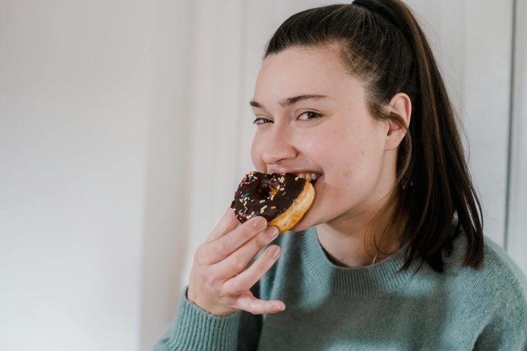 Delighted young female biting delicious sweet chocolate donut and looking at camera while smiling