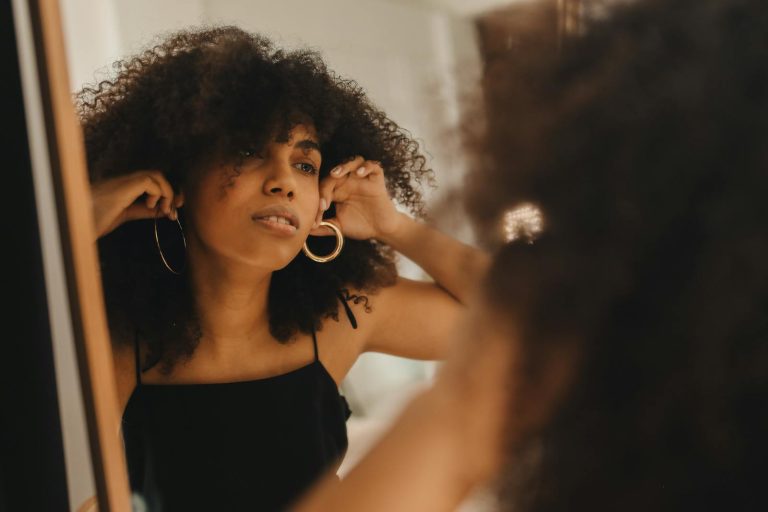 A fashionable woman with curly hair adjusts her earrings in the mirror.