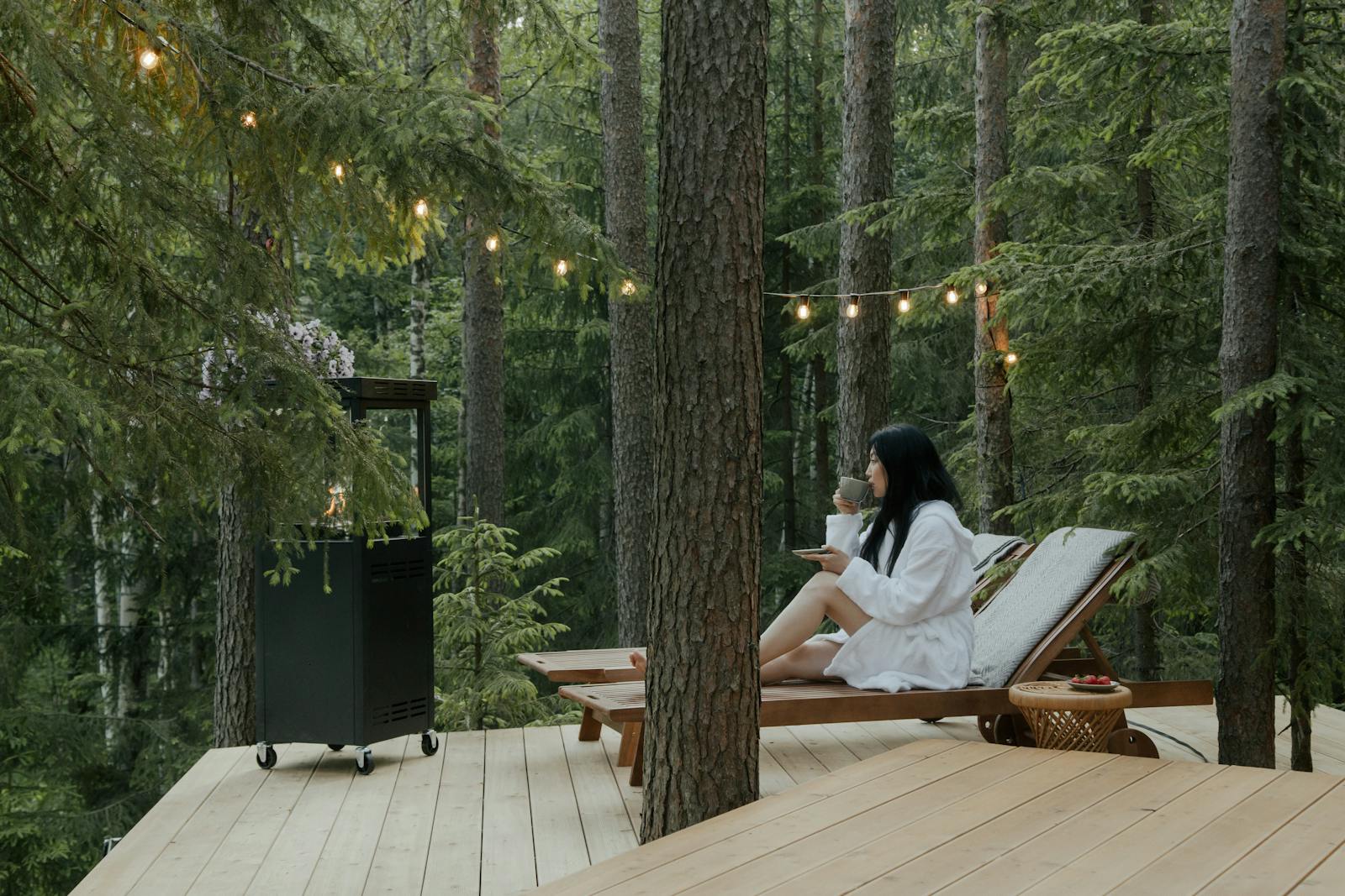Woman in bathrobe relaxing on a deck with drink, surrounded by forest tranquility.