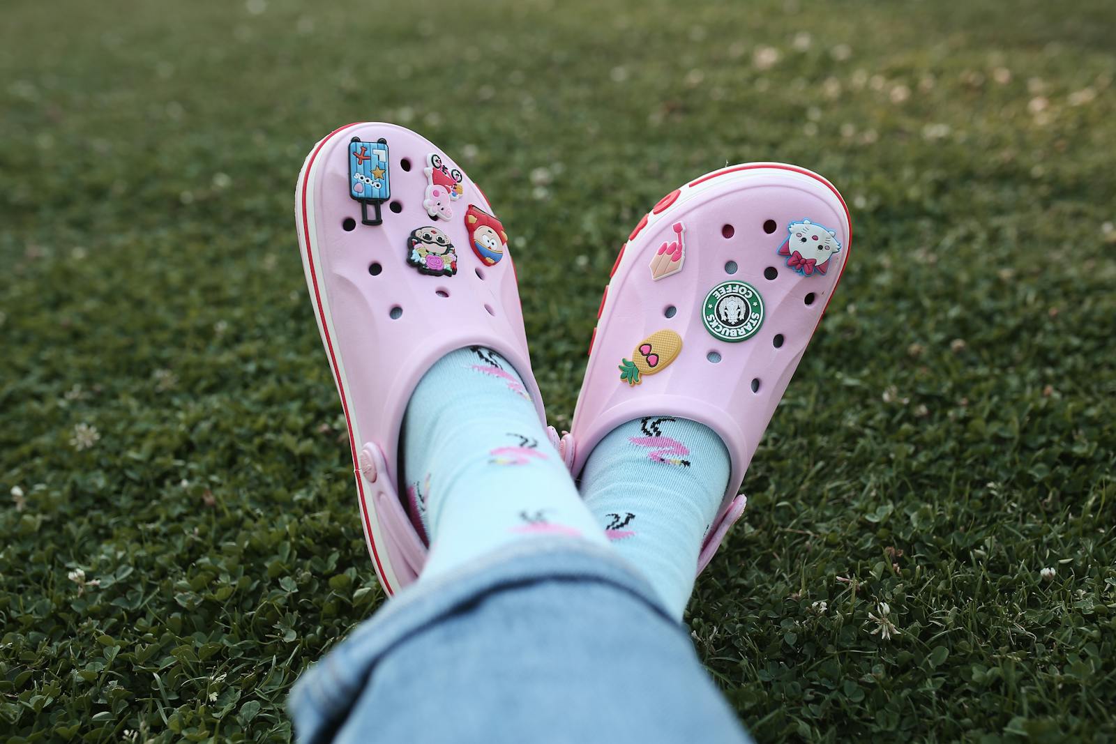 Feet in pink clogs adorned with charms resting on lush grass, showcasing unique style.