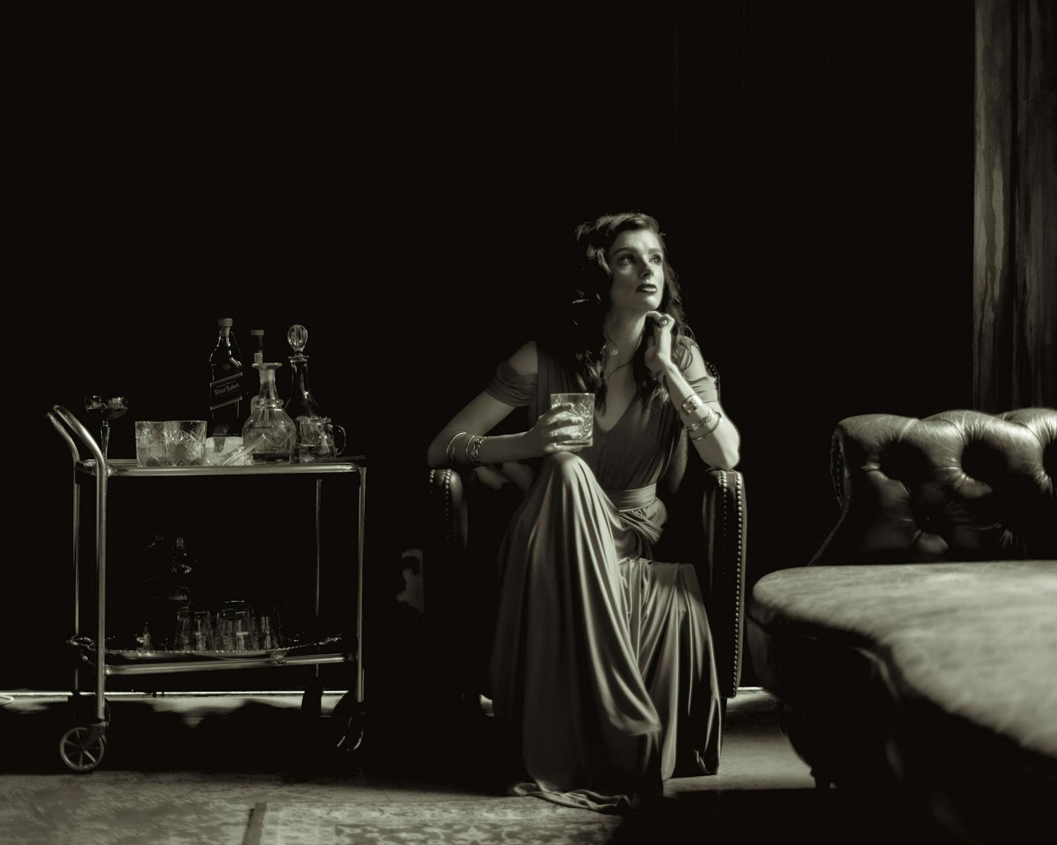 A woman in a flowing dress sits elegantly with a drink in a vintage black and white setting.