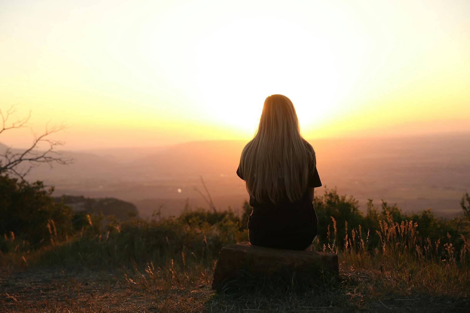 Woman sitting in nature, watching sunset over fields. Peaceful and serene outdoor scene.