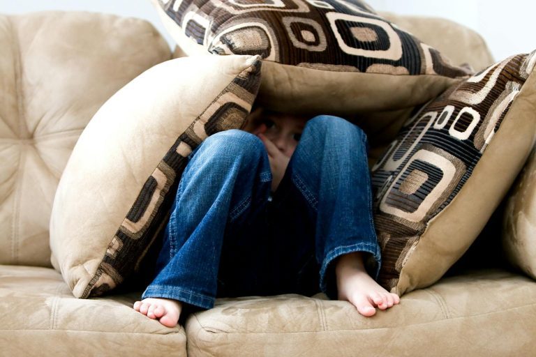 A child crouched under cushions on a couch, conveying a sense of playfulness or fear.