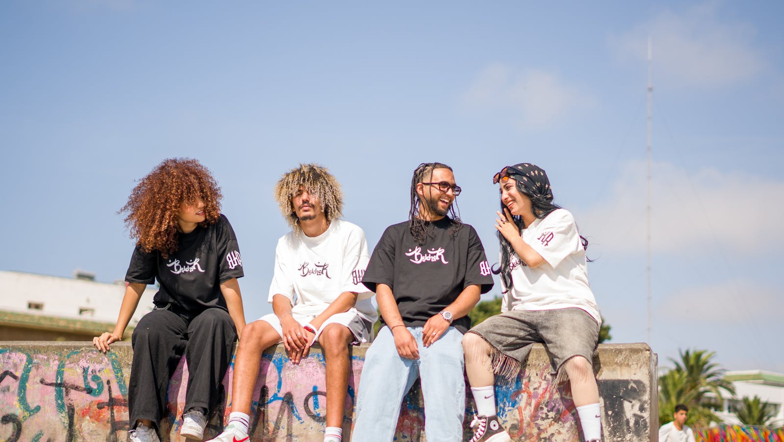 Four friends in casual attire sitting on a graffiti wall, enjoying a sunny day.