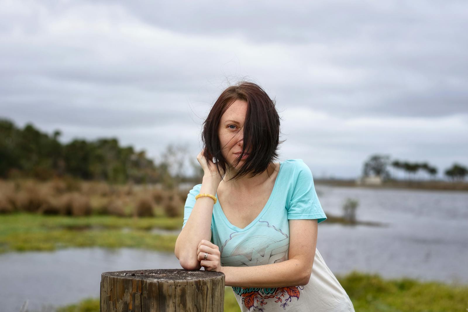 A young woman with windswept hair poses by a tranquil lake under cloudy skies.