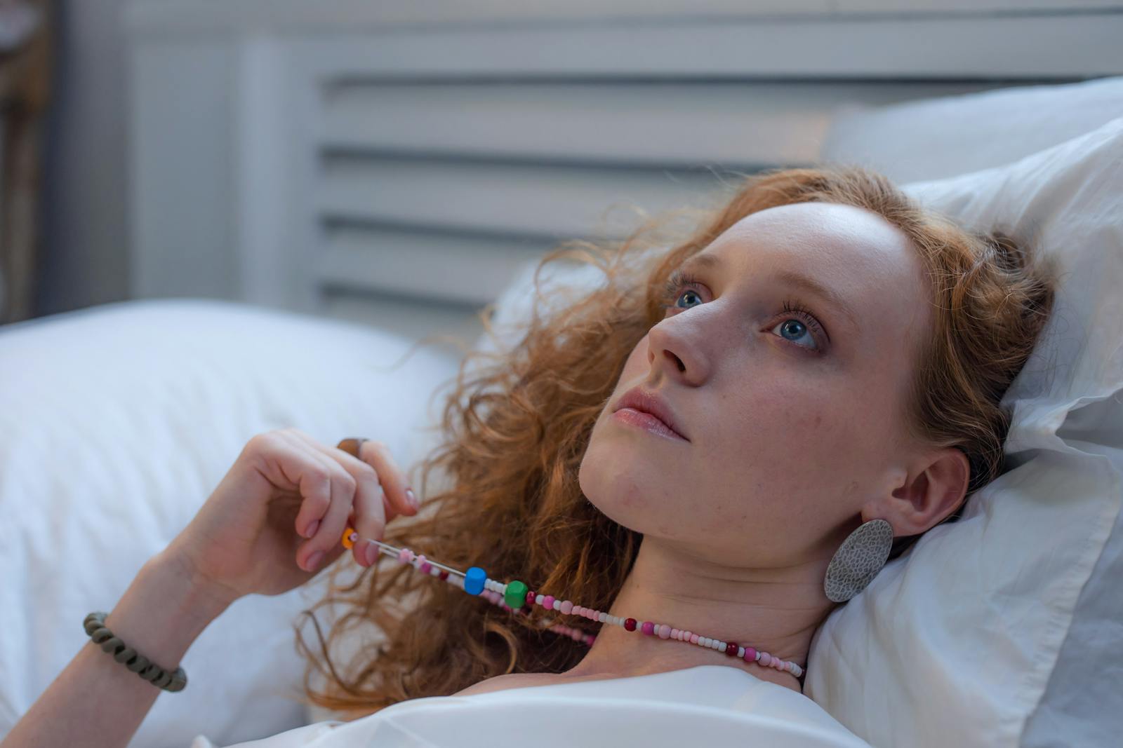 A young redheaded woman lies indoors contemplating while holding a beaded necklace.