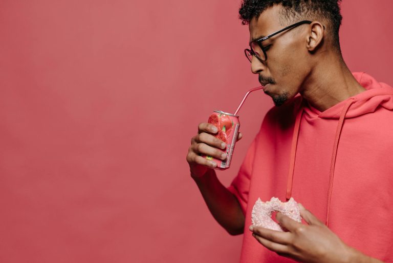 A stylish man drinks soda and holds a donut in a red themed studio setting.