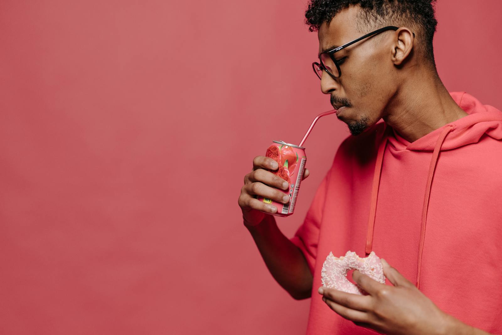 A stylish man drinks soda and holds a donut in a red themed studio setting.