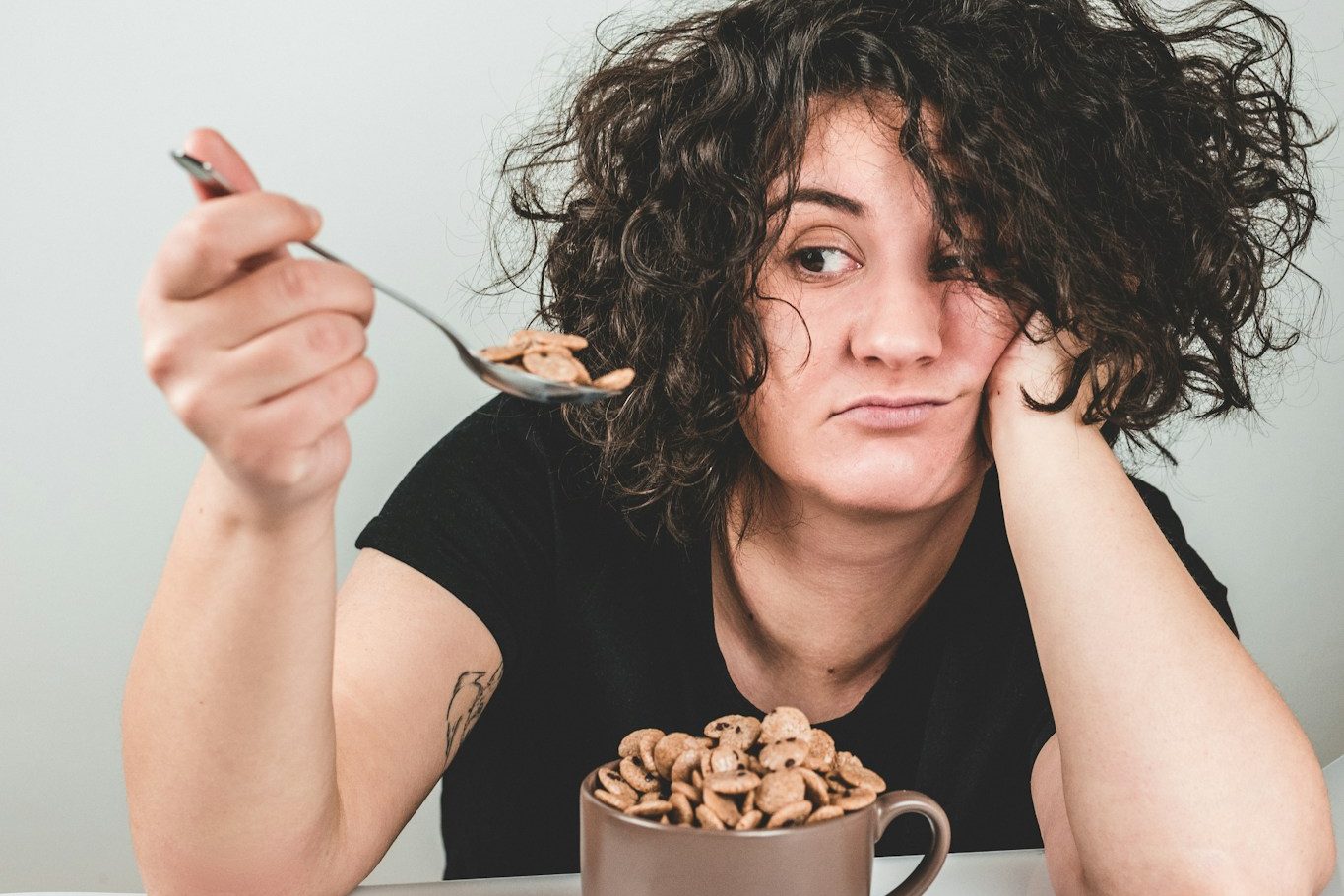 woman with messy hair wearing black crew-neck t-shirt holding spoon with cereals on top