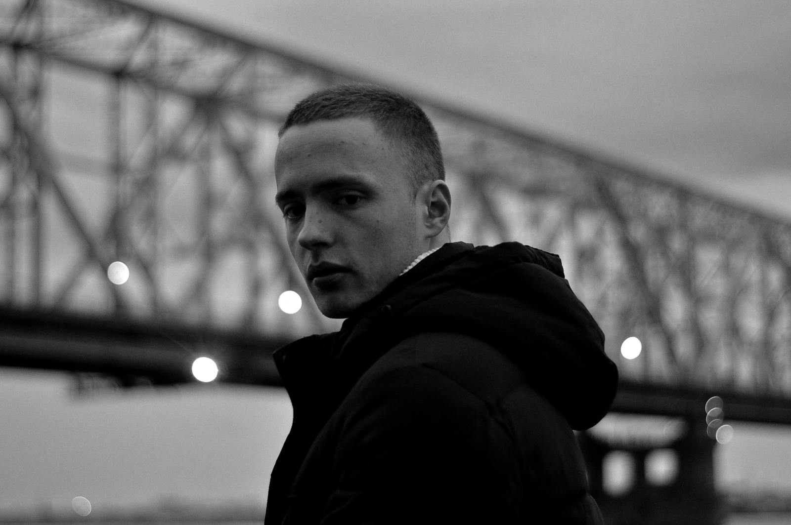 Black and white portrait of a man in a black jacket with a bridge in the background.