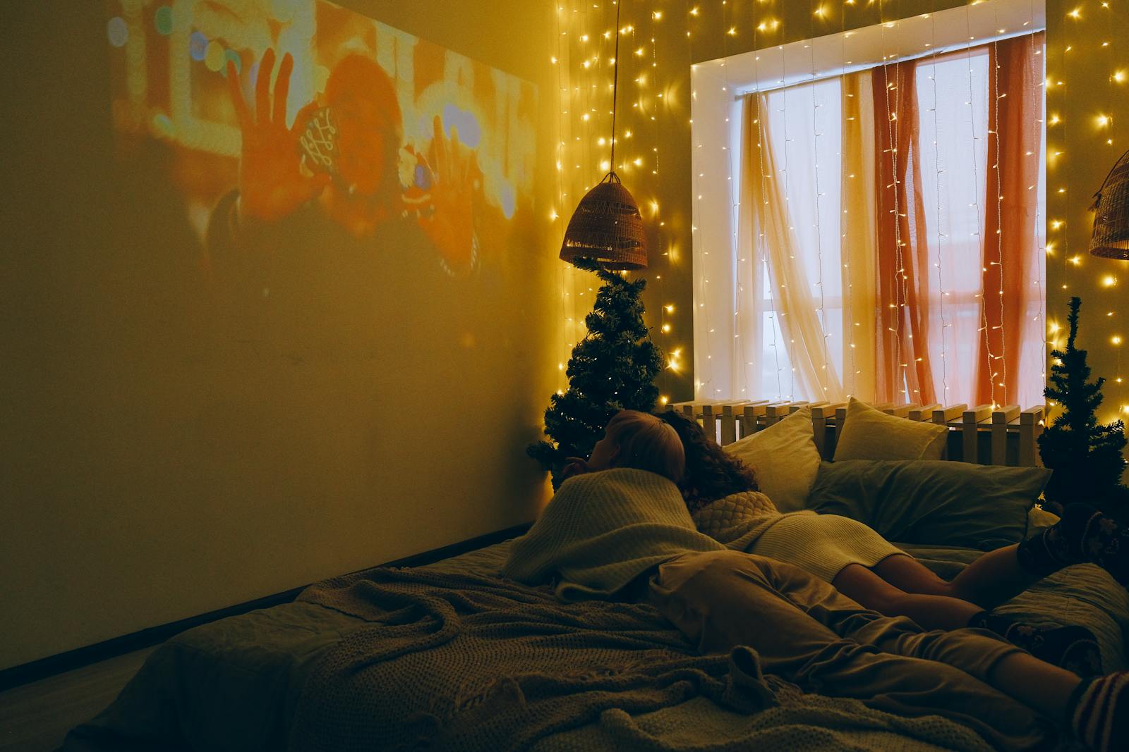 People watching a movie in a warmly lit bedroom decorated with festive lights and small Christmas trees.