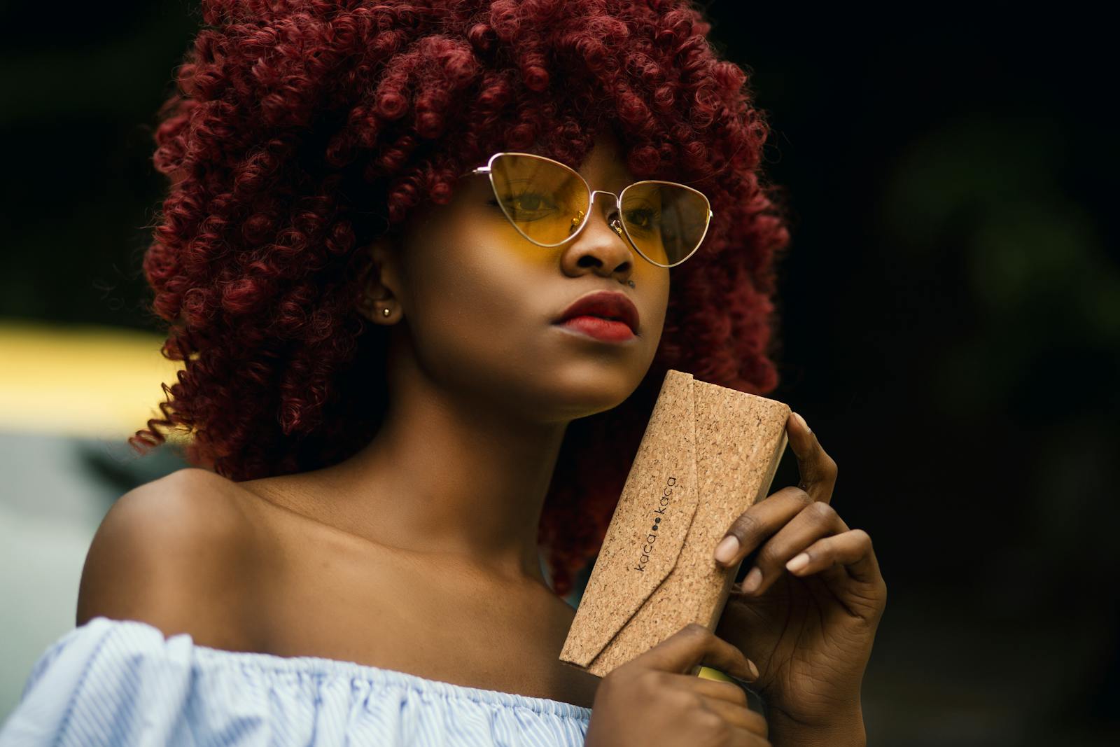 Portrait of a fashionable woman with red curly hair, wearing sunglasses and holding a cork wallet.