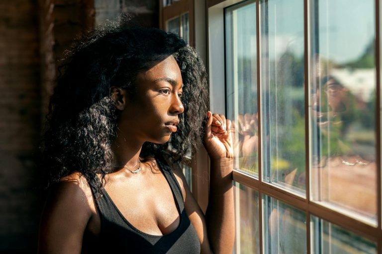 A contemplative woman with curly hair gazes thoughtfully through a sunlit window.