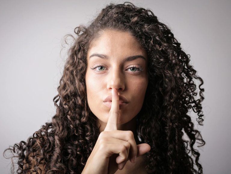 Close-up portrait of a woman with curly hair, signaling quiet with a finger on lips.