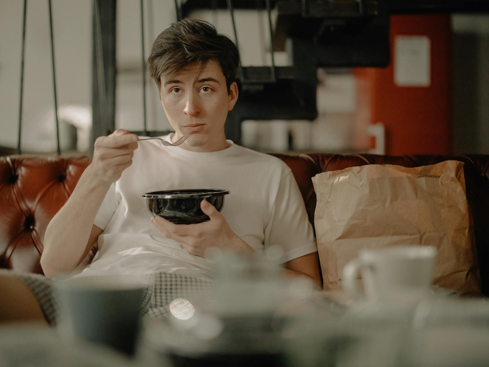 A young man sitting on a couch in an apartment, enjoying his meal with a paper bag nearby.