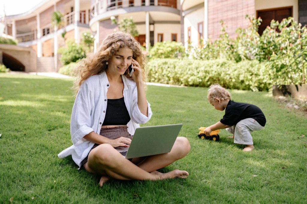 Woman multitasking in garden with laptop and phone, child playing nearby.