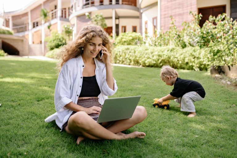 Woman multitasking in garden with laptop and phone, child playing nearby.