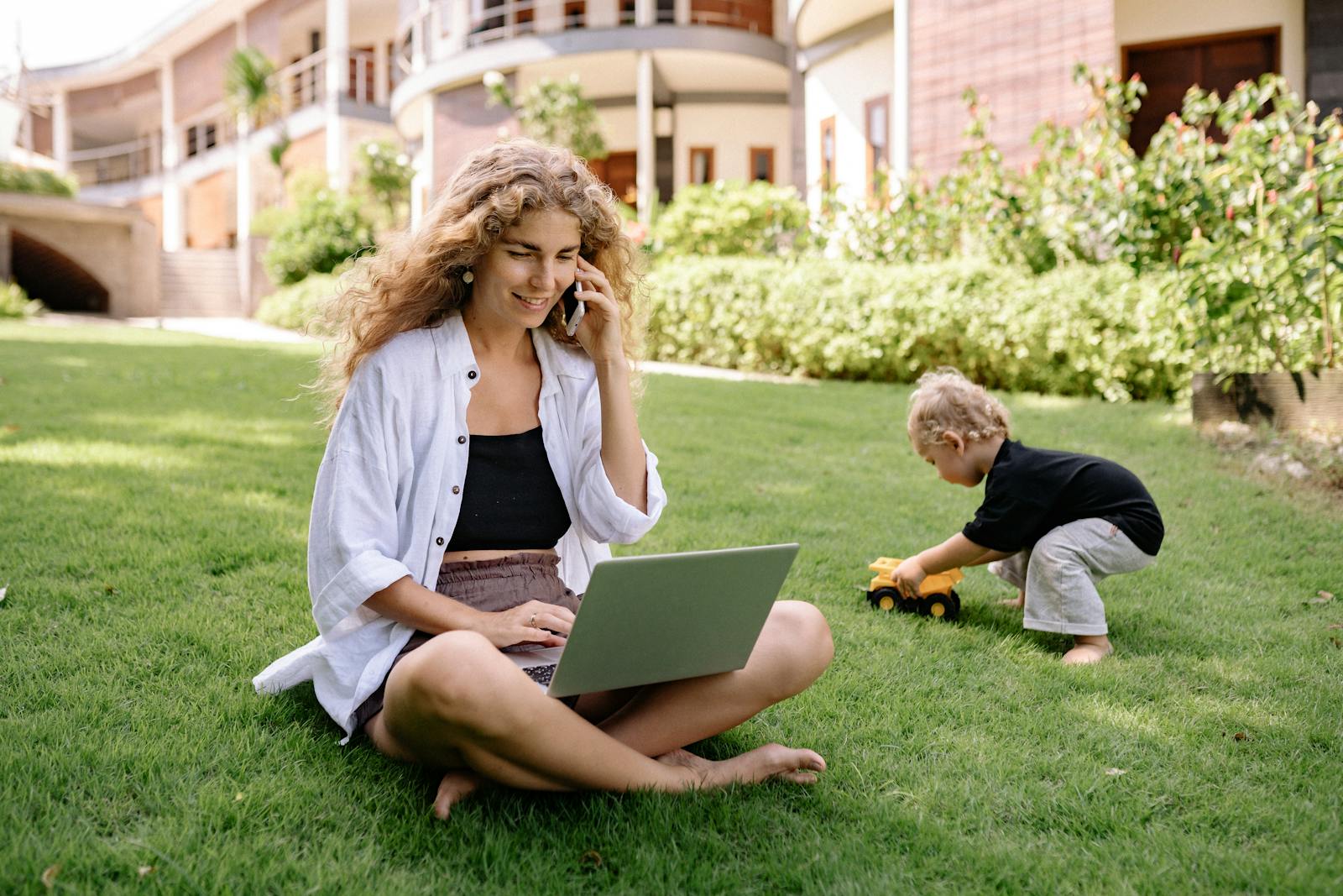 Woman multitasking in garden with laptop and phone, child playing nearby.