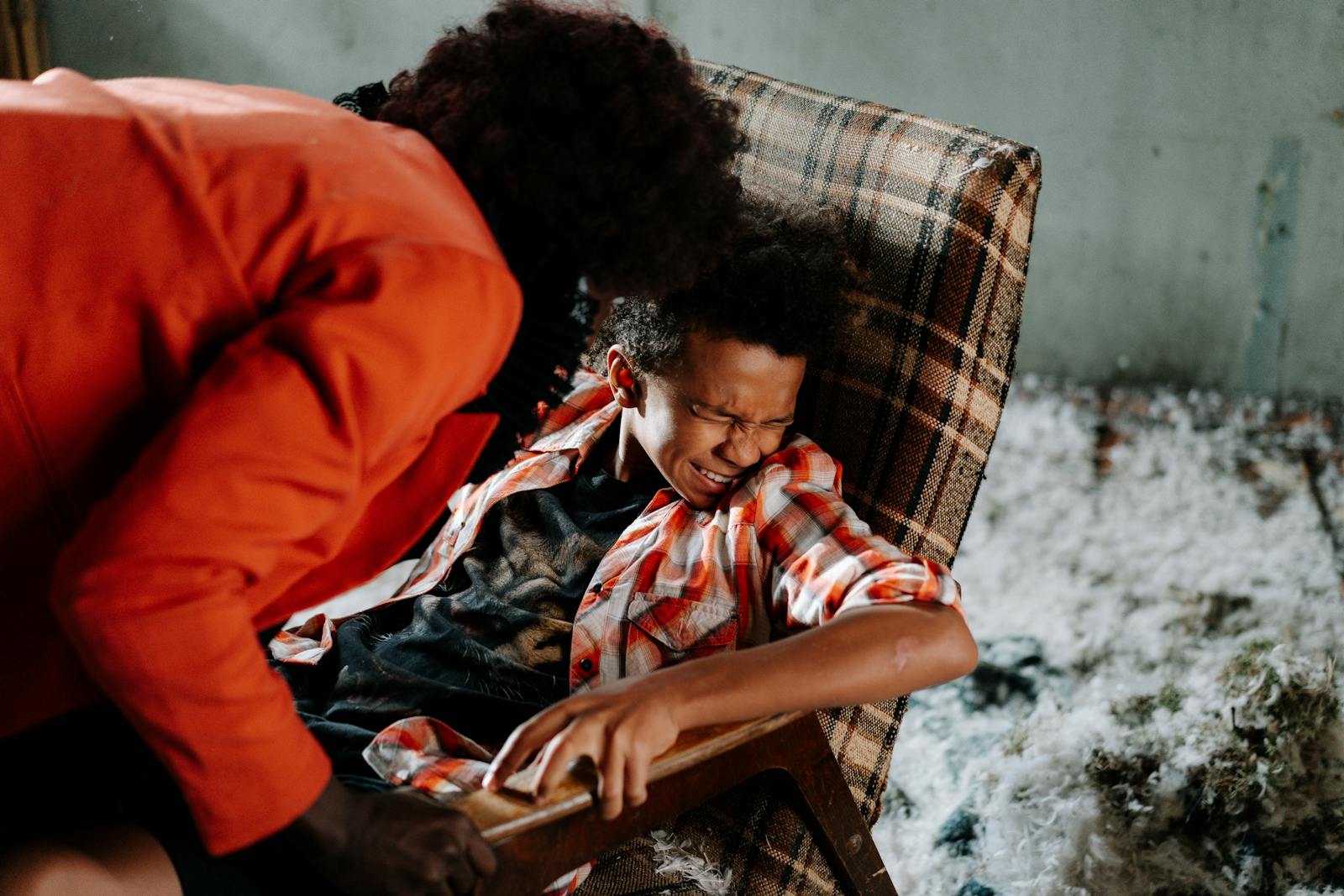 Child in distress on checkered chair with adult leaning over ominously inside a dimly lit room.