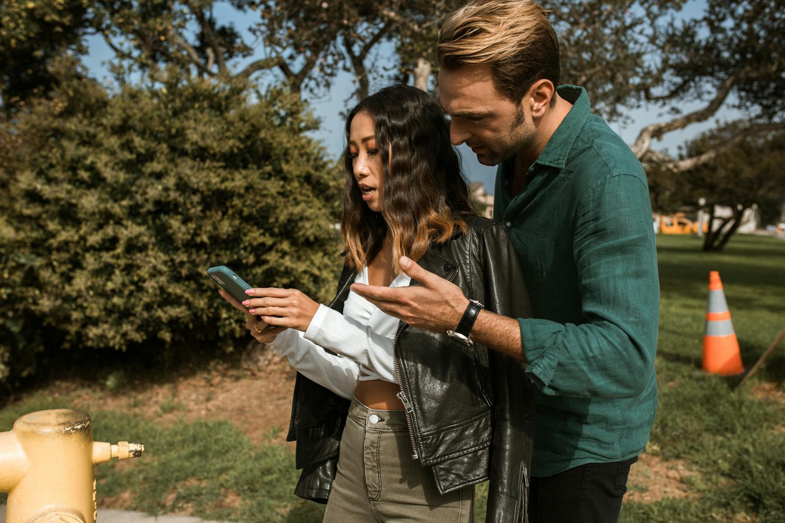 A couple having a disagreement outdoors, focusing on a cellphone. Emotions are visible.