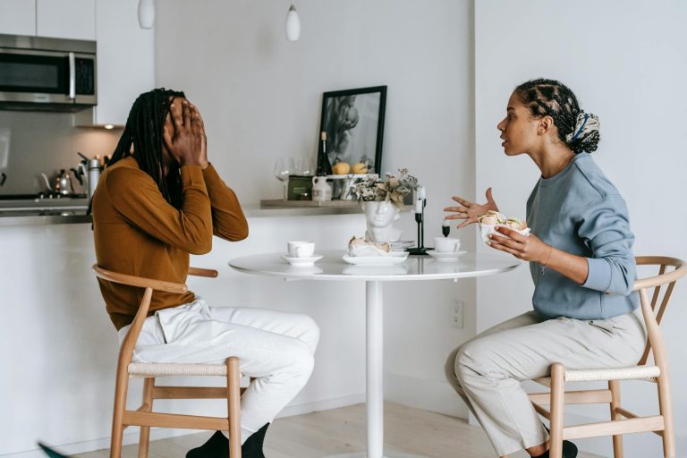 Side view of expressive Hispanic female in casual clothes arguing with African American boyfriend covering face with hands while sitting at table in kitchen at home